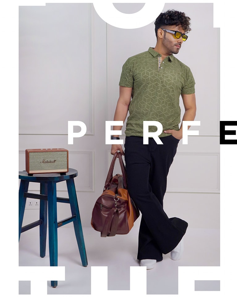 Male model standing next to a blue stool, wearing an olive green patterned polo shirt and holding a brown leather duffel bag, accessorized with yellow-tinted sunglasses.
